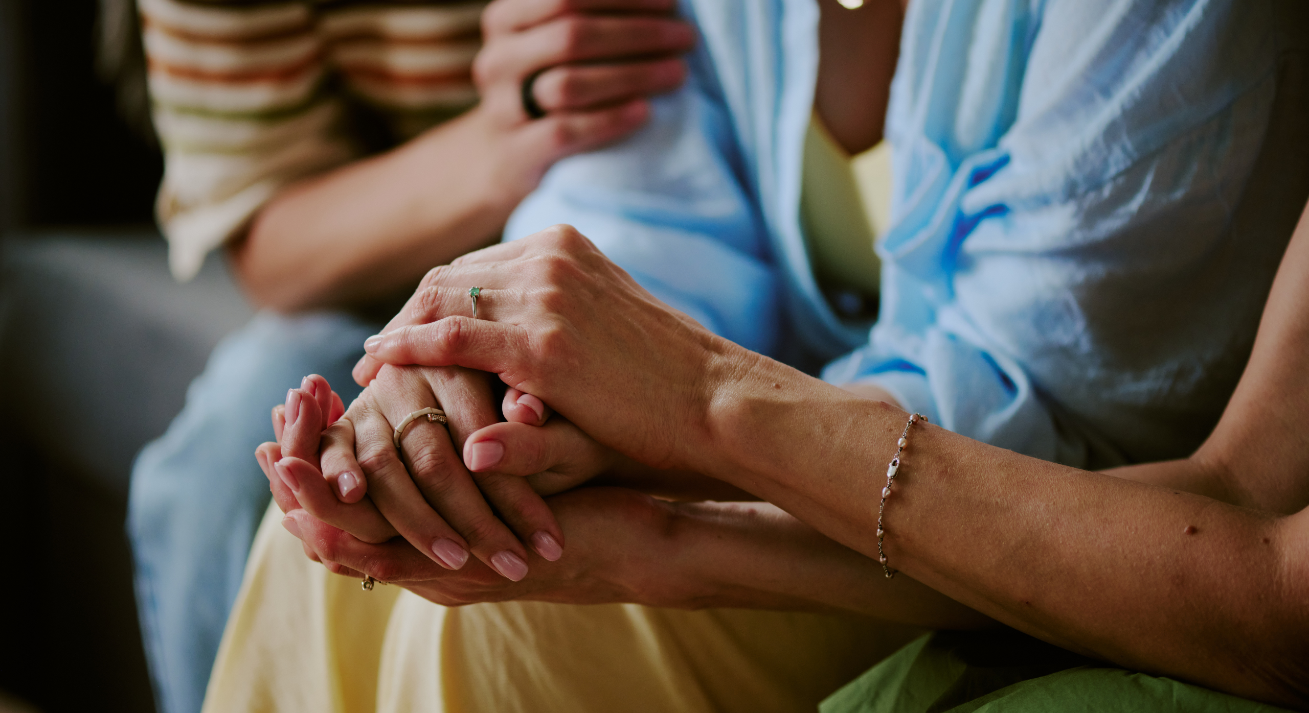 Two pairs of hands clasped together in a comforting grip, with rings and bracelets visible on the wrists—suggesting support.