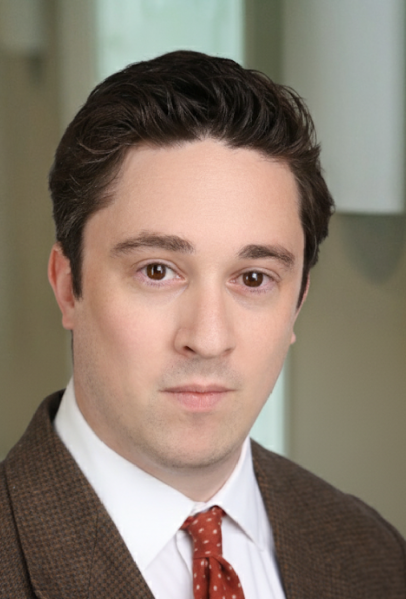 Head-and-shoulders portrait of a man in a dark brown suit and red patterned tie, office background