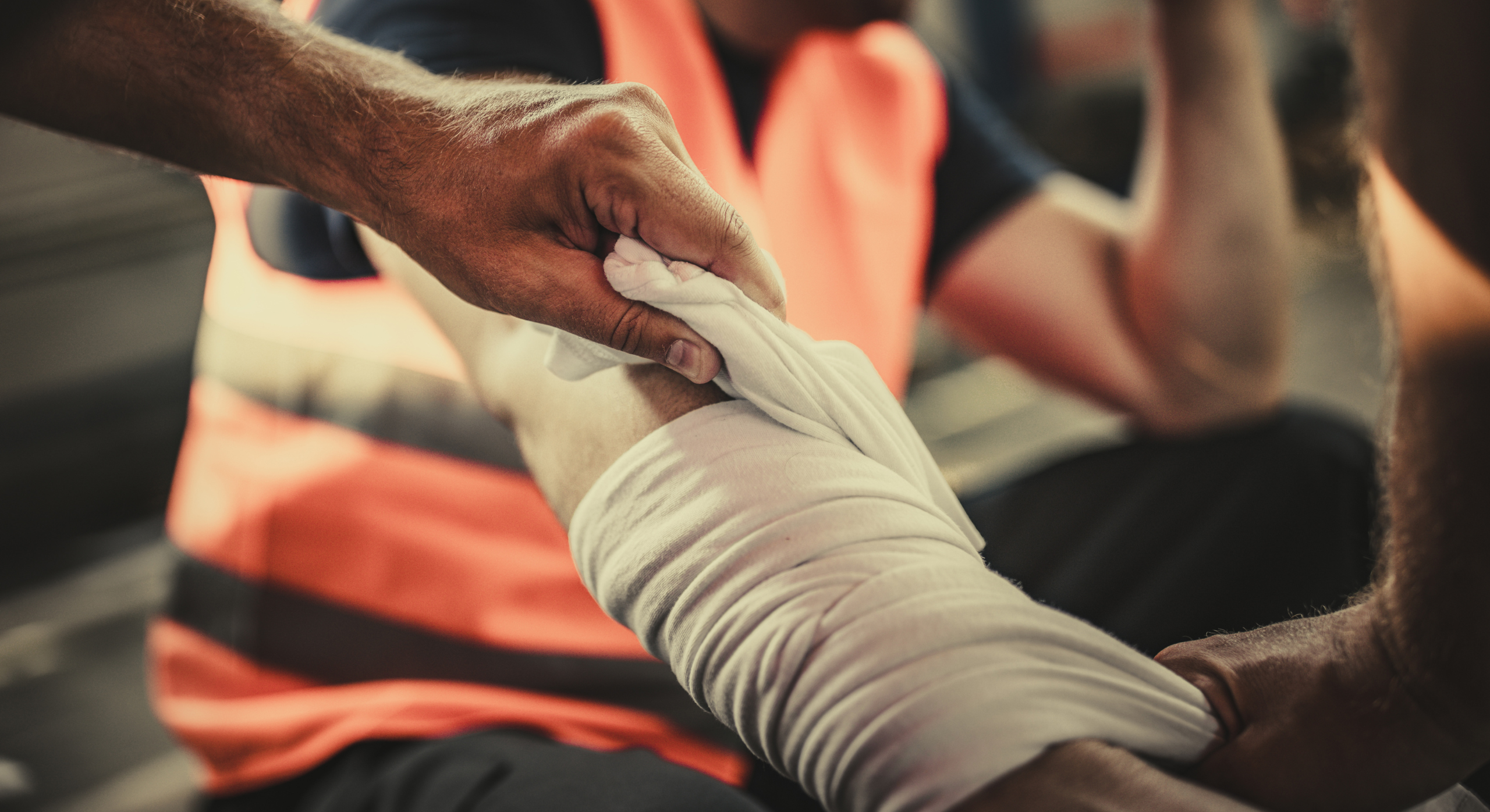 Close-up of a helper wrapping a white cloth bandage around an injured forearm, with an orange safety vest in the background.